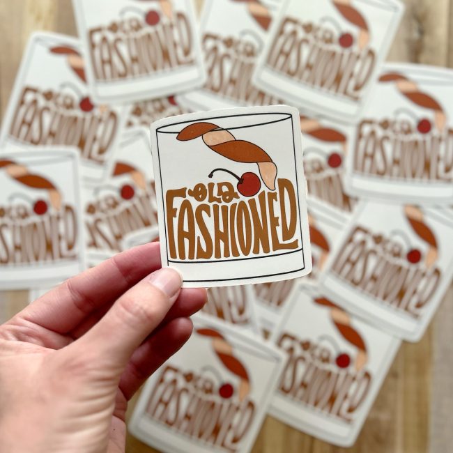A hand holding a "old fashioned" sticker where the bourbon in the glass is actually the lettering which reads "old fashioned." More old fashioned stickers in the background