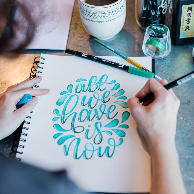 A view over the shoulder of Cortlan hand lettering in her sketchbook which reads "All we have is now" in blue and teal script watercolor lettering.