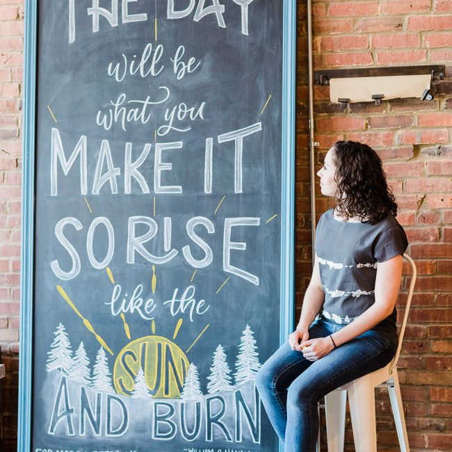 Cortlan sitting next to her large scale chalkboard lettering that reads "The Day will be What You Make it so Rise like the Sun and Burn"