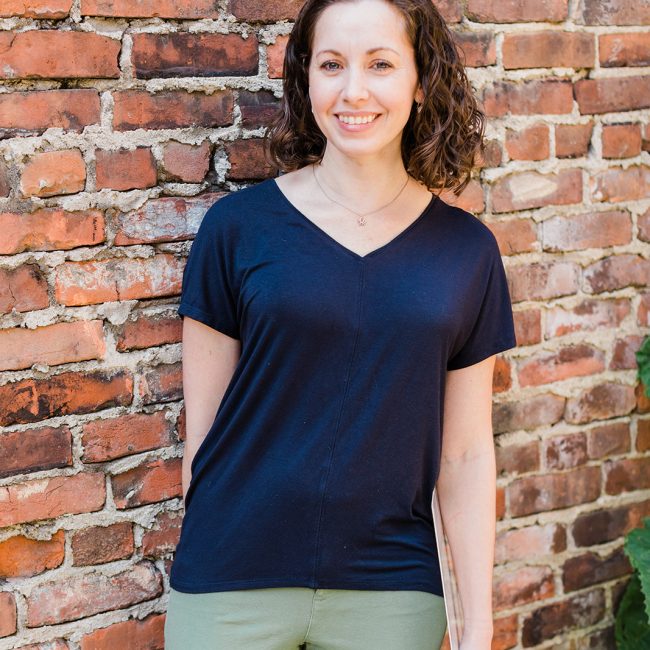 A portrait of Cortlan wearing navy blue with pale green pants in front of a brick wall.